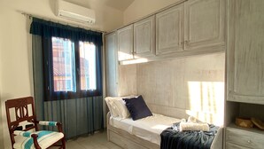 A single bed with a headboard, a wooden chair with a cushion, a window with curtains, and a wall-mounted air conditioner.