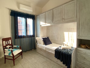 A single bed with a headboard, a wooden chair with a cushion, a window with curtains, and a wall-mounted air conditioner.
