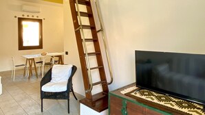 A living room with a wooden ladder against the wall, a TV on a wooden cabinet, a dining area with chairs and a table, and a staircase.