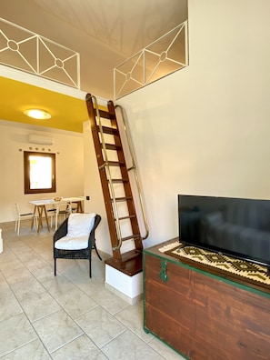 A living room with a wooden ladder against the wall, a TV on a wooden cabinet, a dining area with chairs and a table, and a staircase.