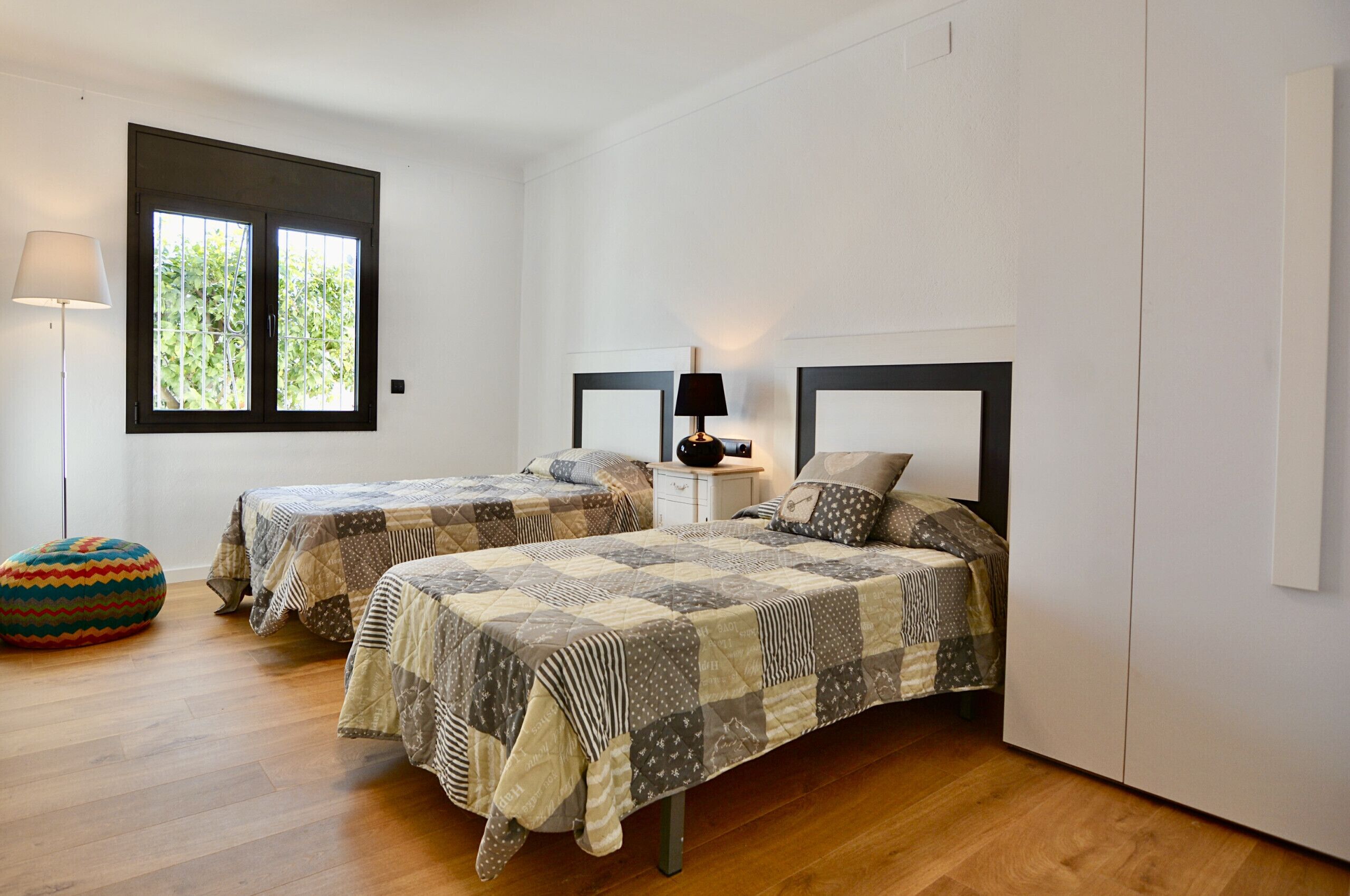 A bedroom with two beds, a window, a lamp, and a colorful ottoman.
