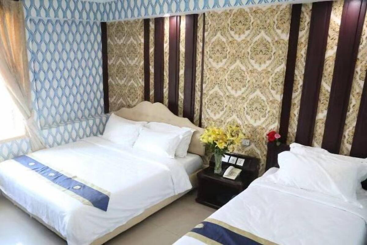 A hotel room with a large bed, a smaller bed, a nightstand with a vase of flowers, and patterned wallpaper.