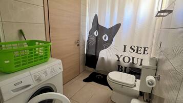 A compact bathroom with a washing machine, toilet, and a shower curtain featuring a cat design.