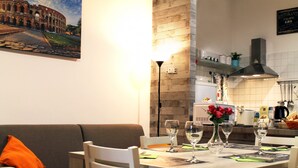 A dining area with a table set for two, a sofa with an orange pillow, and a wall-mounted picture of the Colosseum. - 50 Steps Colosseum Apartment (Roma)