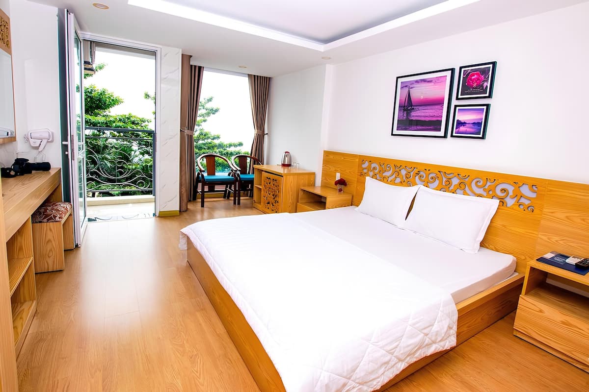 Une chambre d’hôtel avec un grand lit, une tête de lit en bois, un bureau avec une chaise et un balcon donnant sur un paysage verdoyant.