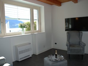 Una sala de estar moderna con un gran ventanal, un televisor de pantalla plana montado en la pared, un sillón gris y una mesita con una botella de champán.