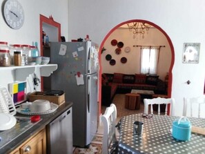 A compact kitchen with a refrigerator, a dining table with chairs, and a living area with a sofa and a TV. - Houch La Driba (Djerba Midun)