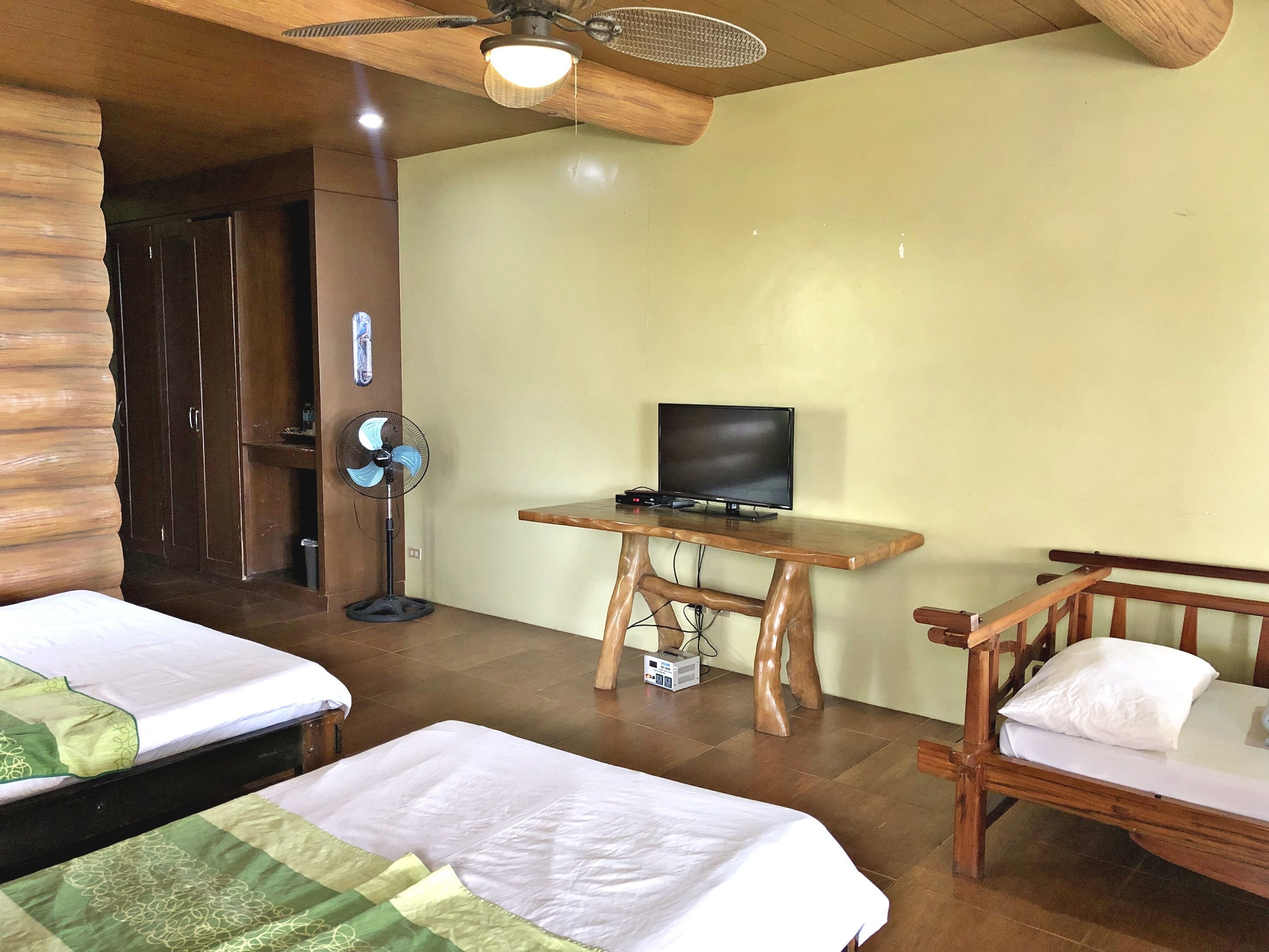 Une chambre avec deux lits, un bureau en bois avec une télévision, un ventilateur et un ventilateur de plafond.