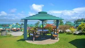 A gazebo with a green roof and wooden benches, surrounded by greenery and a clear sky.