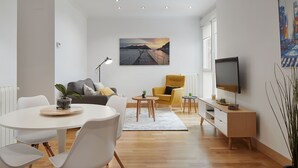 Una sala de estar moderna con comedor, un sofá, un sillón amarillo, un televisor y un cuadro de una escena costera.