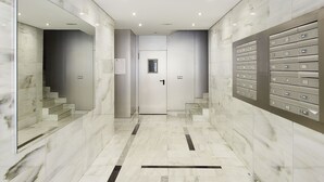 Un pasillo moderno con suelo de mármol, una puerta con un pequeño ventanal y una fila de taquillas metálicas a la derecha.