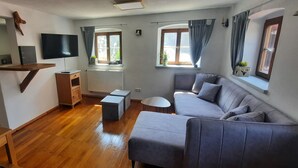 A compact living space with a kitchenette, a grey sofa, a small table, and a TV mounted on the wall. - Vacation home on the romantic road (Landsberg/Erpfting)