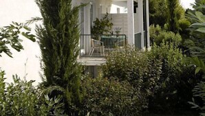 Un edificio de dos pisos con balcones, rodeado de abundante vegetación y un pequeño elemento acuático.