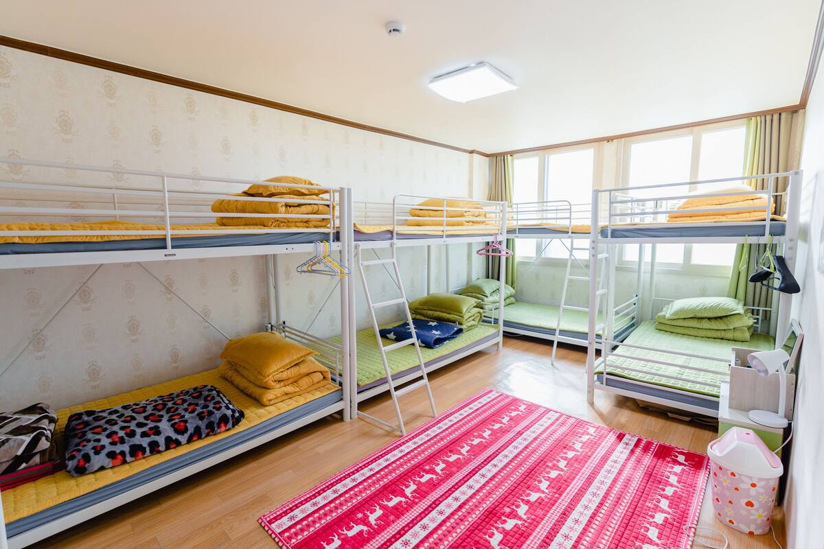 A dormitory room with bunk beds, a red patterned rug, and a small desk with a lamp.