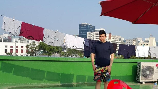 Una terraza en la azotea con suelo verde, sillas rojas y una mesa. Se ven prendas de vestir colgadas en un tendedero al fondo.