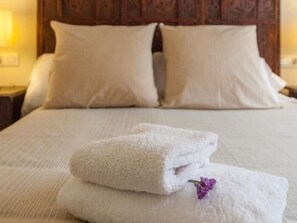 Cama bem arrumada com lençóis e almofadas brancas, uma toalha branca dobrada com uma flor roxa e uma cabeceira de madeira.