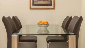 A dining table with a glass top and four chairs, a bowl of fruit on the table, and a framed picture on the wall.