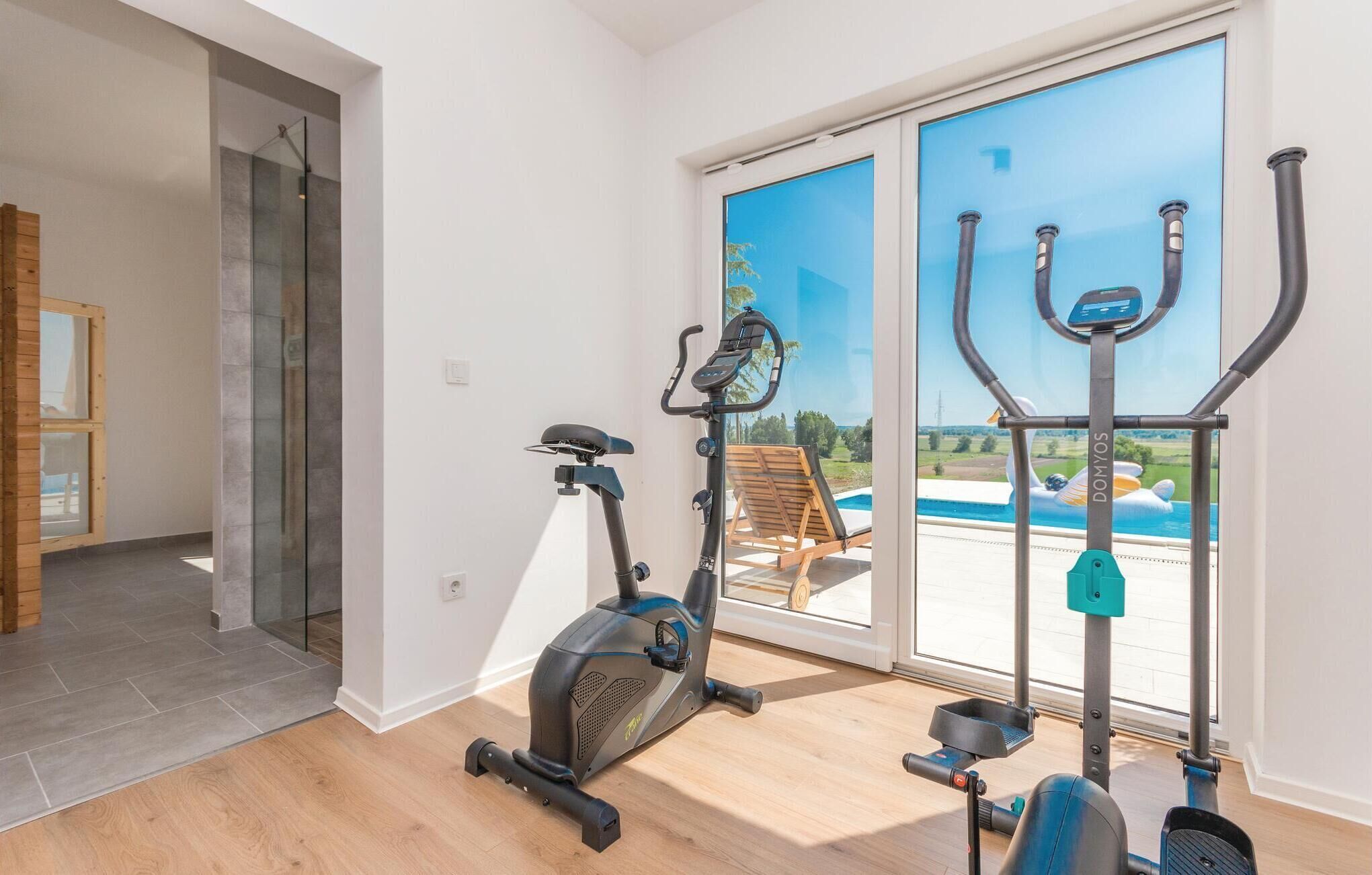Ein Heimfitnessraum mit Ergometer und Crosstrainer, Holzboden und Blick auf ein Schwimmbecken durch große Fenster.