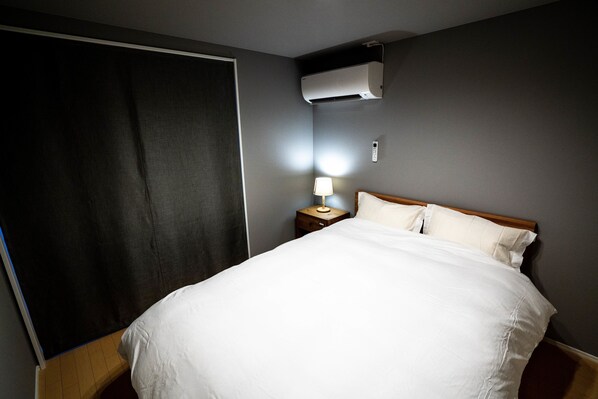 A hotel room with a bed, a nightstand with a lamp, an air conditioning unit, and a dark curtain.