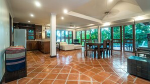 A spacious living area with a kitchen, dining area, and large windows offering views of greenery. - Nakatani Villas By Lofty (Kamala)