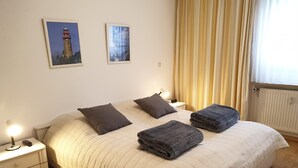 A bedroom with a large bed, two bedside tables, a nightstand, a lamp, and two framed pictures on the wall. - Vacation apartment Parkpension W06 (Laboe)