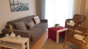 A living room with a sofa, a small table, a wicker chair, a red side table, and a painting on the wall. - Vacation apartment Parkpension W06 (Laboe)