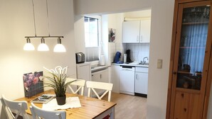 A compact kitchen with white cabinets, a wooden dining table with chairs, and a potted plant. - Vacation apartment Parkpension W06 (Laboe)