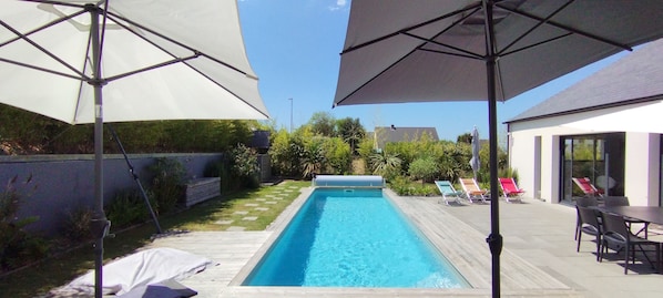 A rectangular swimming pool with a wooden deck surrounding it, two umbrellas, and a patio area with a table and chairs.