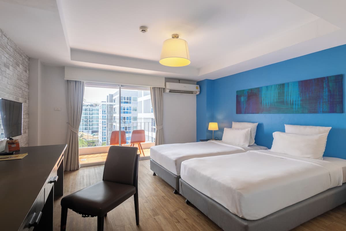 Ein Hotelzimmer mit zwei Betten, einem Schreibtisch, einem Stuhl, einem großen Fenster mit Vorhängen, einer Klimaanlage und einem Bild an der Wand.