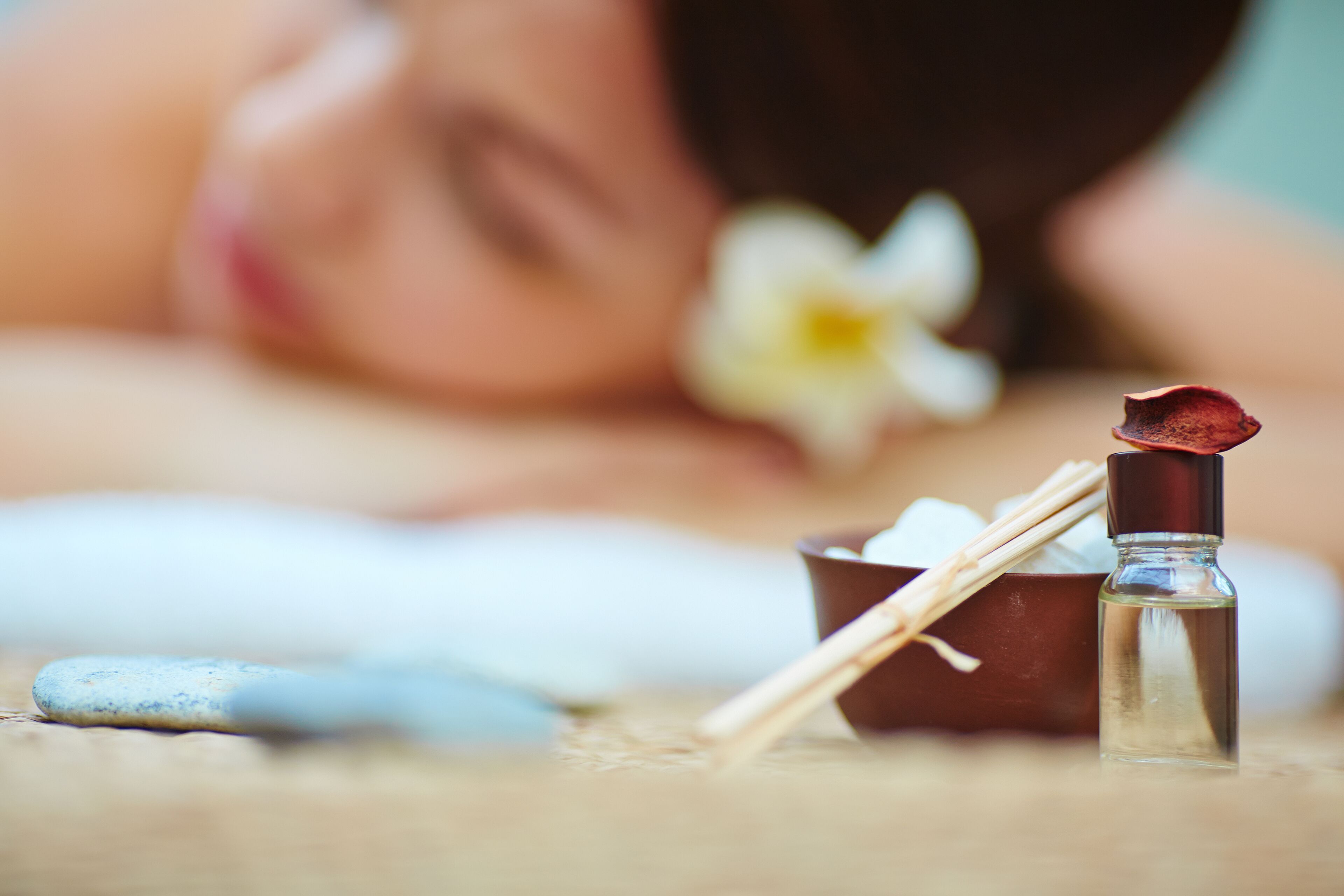 Une scène de spa avec une personne allongée, une petite bouteille d’huile et un vase rempli de fleurs blanches.