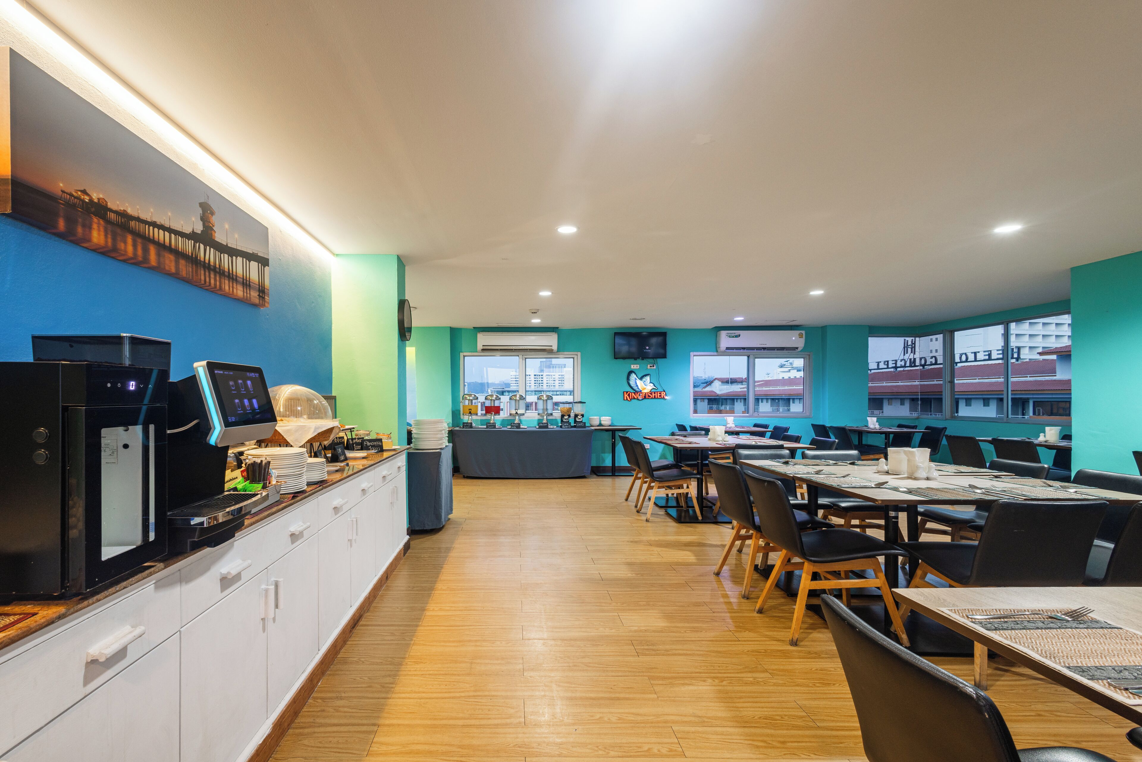 Un espace de repas moderne avec des tables et des chaises, un comptoir de cuisine équipé d’appareils électroménagers et une vue sur les bâtiments à travers de grandes fenêtres.
