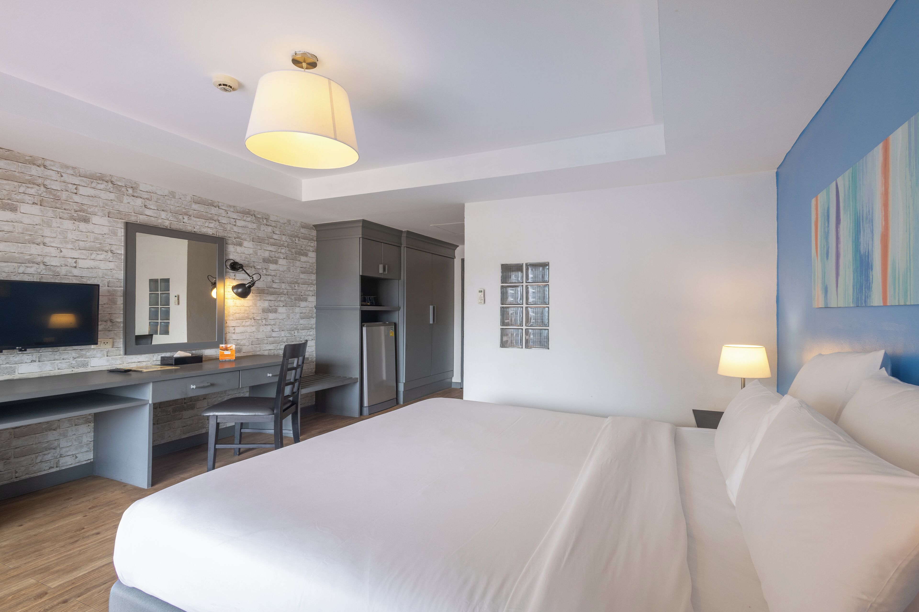 A modern hotel room with a large bed, a desk with a chair, a television, and a kitchenette.