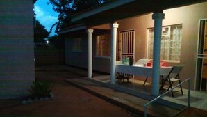 A covered outdoor area with a table and chairs, a railing, and a building with windows and a door. - BJ&T Vacation Homes (Kasane)