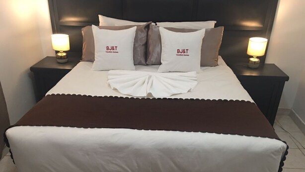 A neatly made bed with a brown and white bedspread, two pillows with "BJ&T Vacation Homes" branding, and two bedside lamps on dark nightstands. - BJ&T Vacation Homes (Kasane)