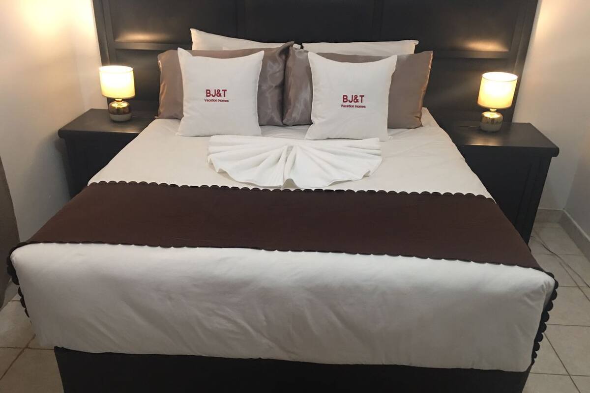 A neatly made bed with a brown and white bedspread, two pillows with "BJ&T Vacation Homes" branding, and two bedside lamps on dark nightstands.