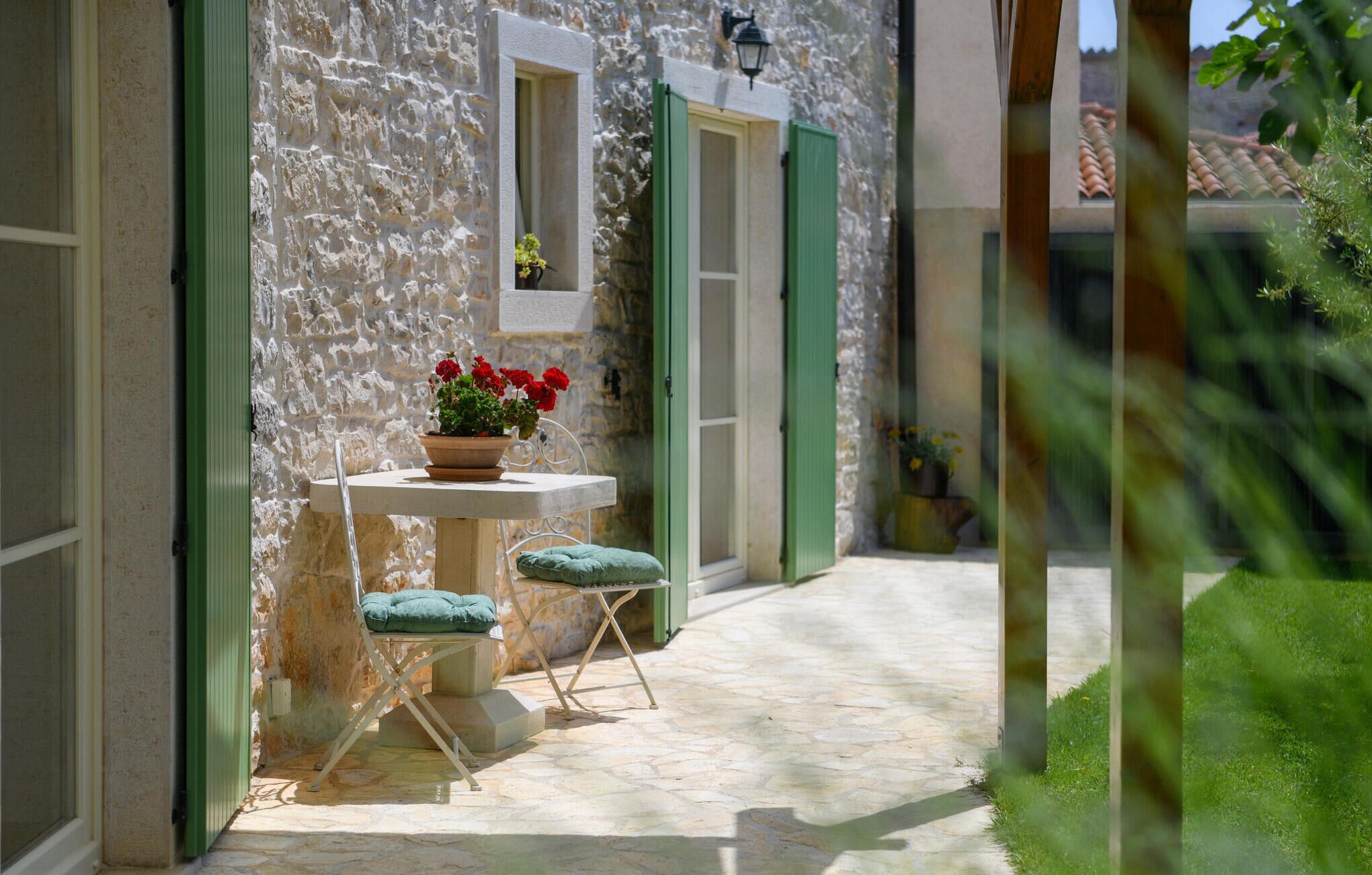 Un bâtiment en pierre avec une petite table et deux chaises, une fenêtre avec des volets verts et un pot de fleurs contenant des fleurs rouges.