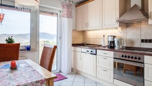 Une cuisine équipée d’une cuisinière, d’un four et d’un évier. On y trouve une table à manger recouverte d’une nappe à motifs floraux et une chaise. Une fenêtre donne sur l’extérieur et offre une vue sur le paysage.