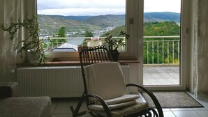 Une pièce avec vue sur une vallée et des montagnes, grâce à une grande fenêtre. Un fauteuil et un pot de fleurs sont posés sur le rebord de la fenêtre.