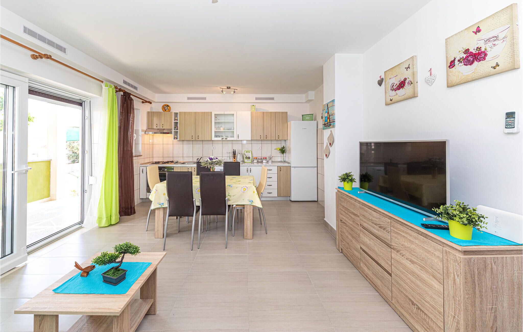 A modern kitchen with wooden cabinets, a dining area with a table and chairs, a TV on a wooden console, and a small potted plant on the console.