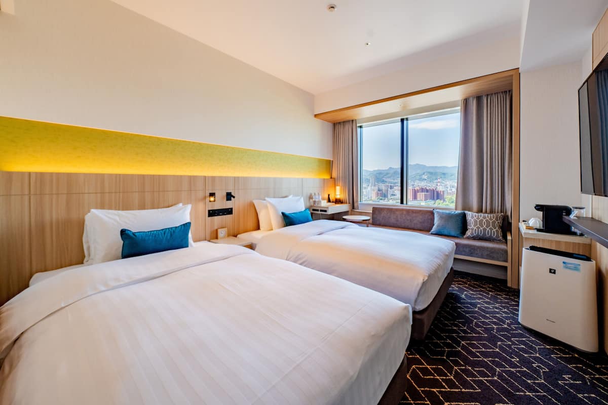 Habitación de hotel con dos camas, un sofá, un escritorio y un amplio ventanal con vista a la ciudad.