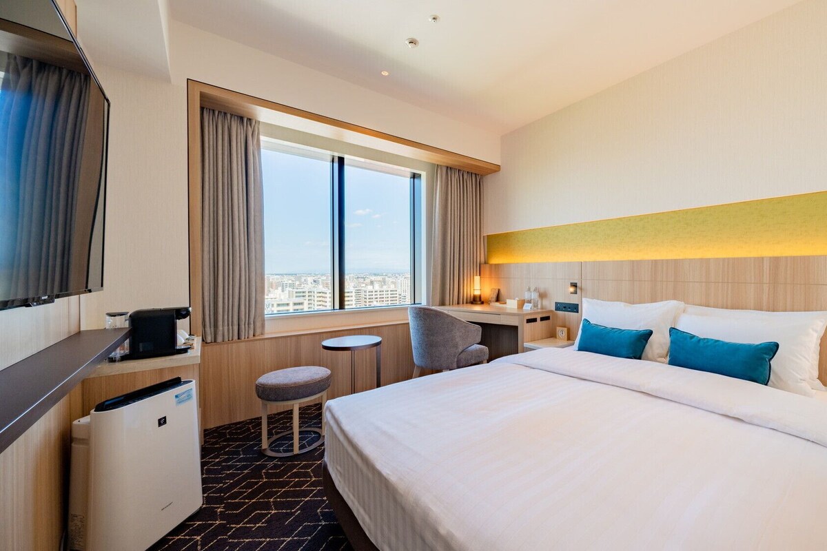 A hotel room with a bed, a desk, a chair, a TV, and a window with a city view.