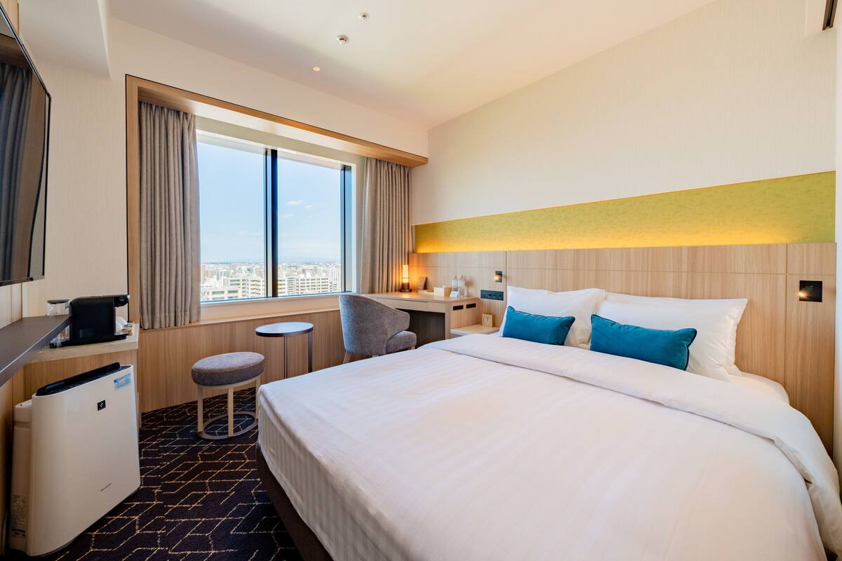 Habitación de hotel con una cama grande, un escritorio, una silla, una mesita y vistas a la ciudad desde la ventana.