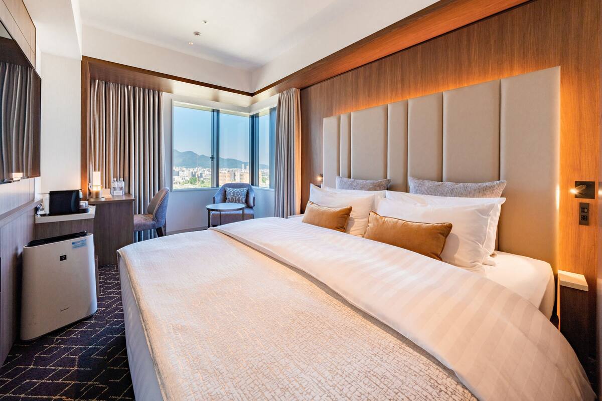 Habitación de hotel con una cama grande, un escritorio con computadora, una silla y vistas de la ciudad a través de amplios ventanales.