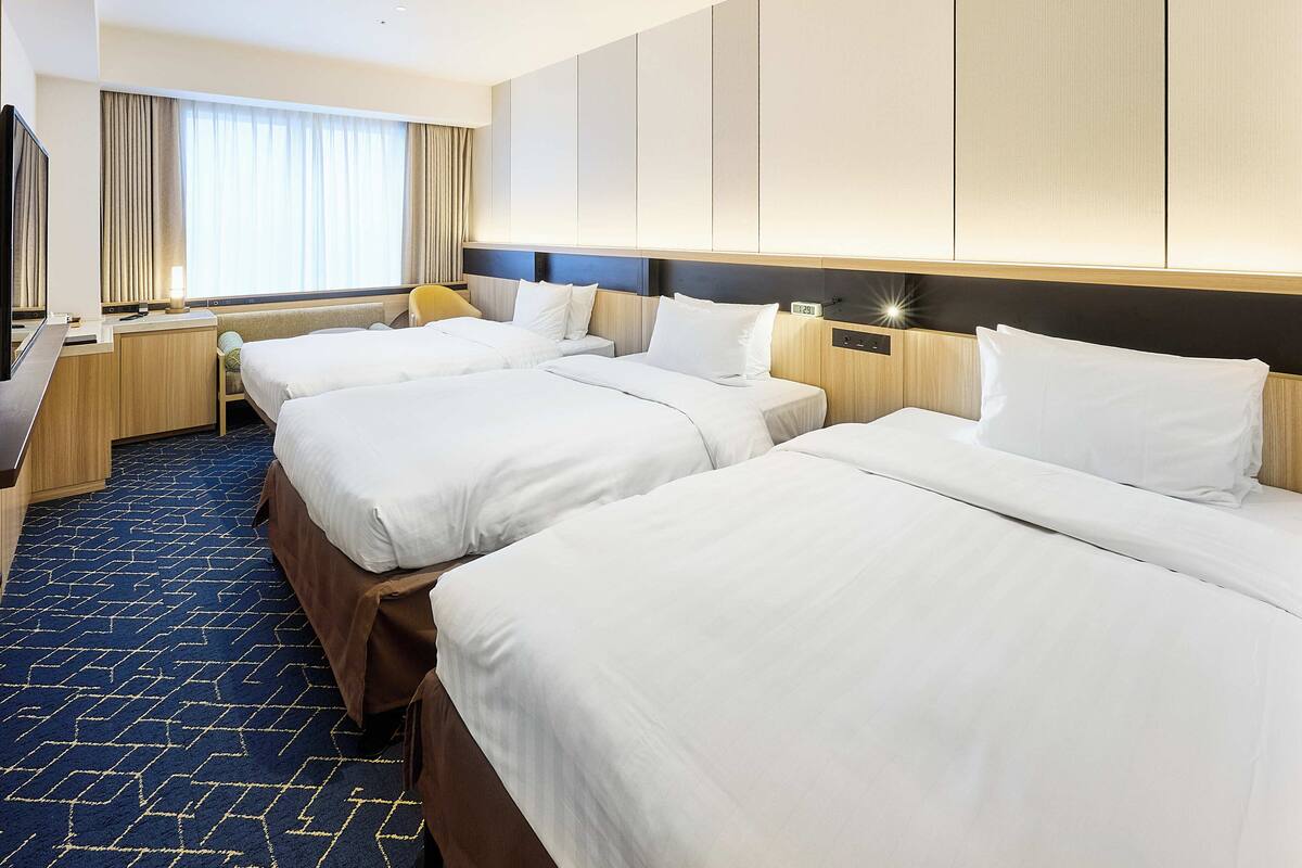 Habitación de hotel con tres camas, un escritorio, una silla y un amplio ventanal con cortinas.