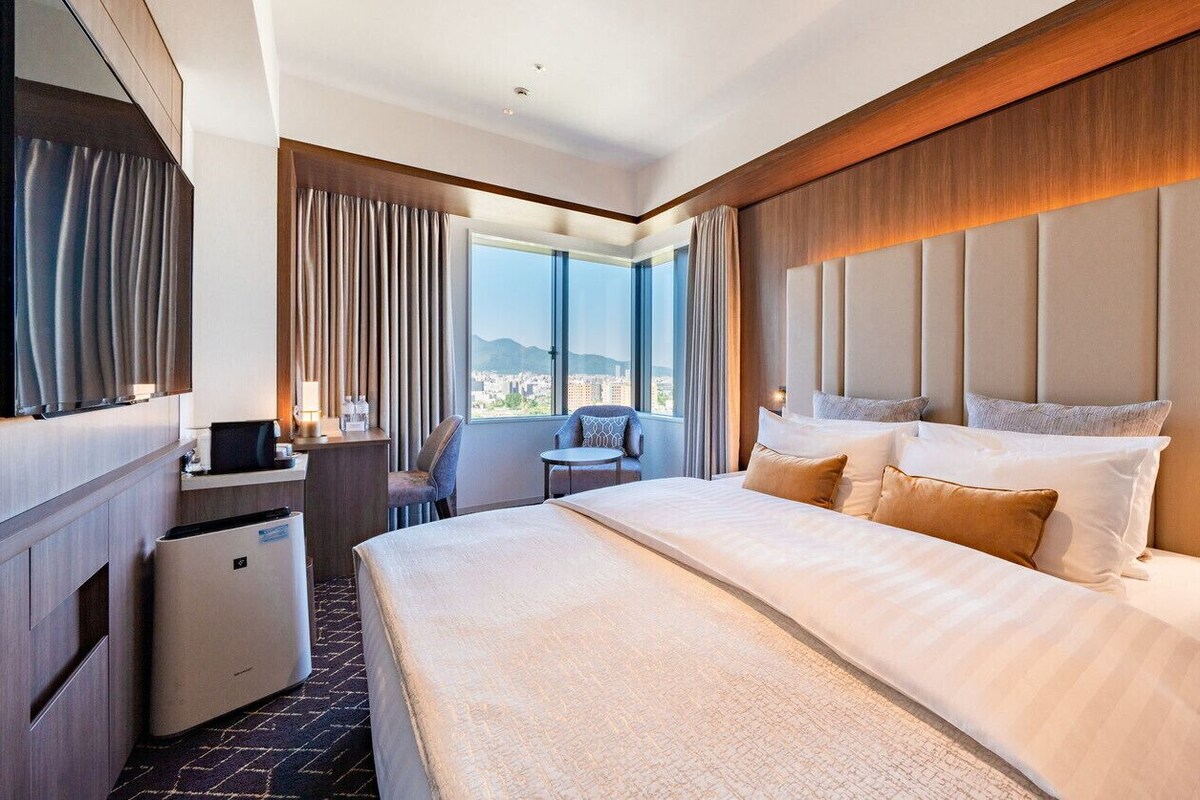 A hotel room with a large bed, a desk, a chair, a small table, and a view of the city through large windows.
