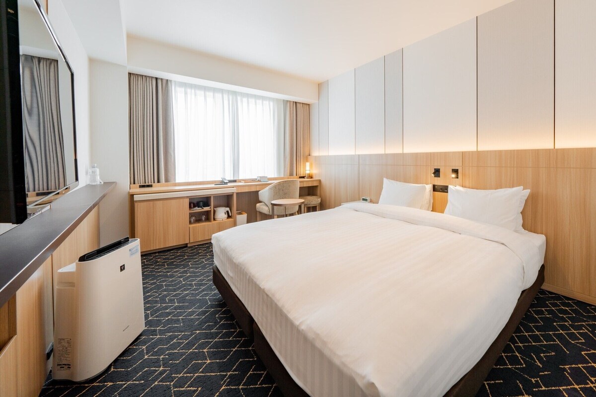 A hotel room with a bed, desk, chair, and a large window with curtains.
