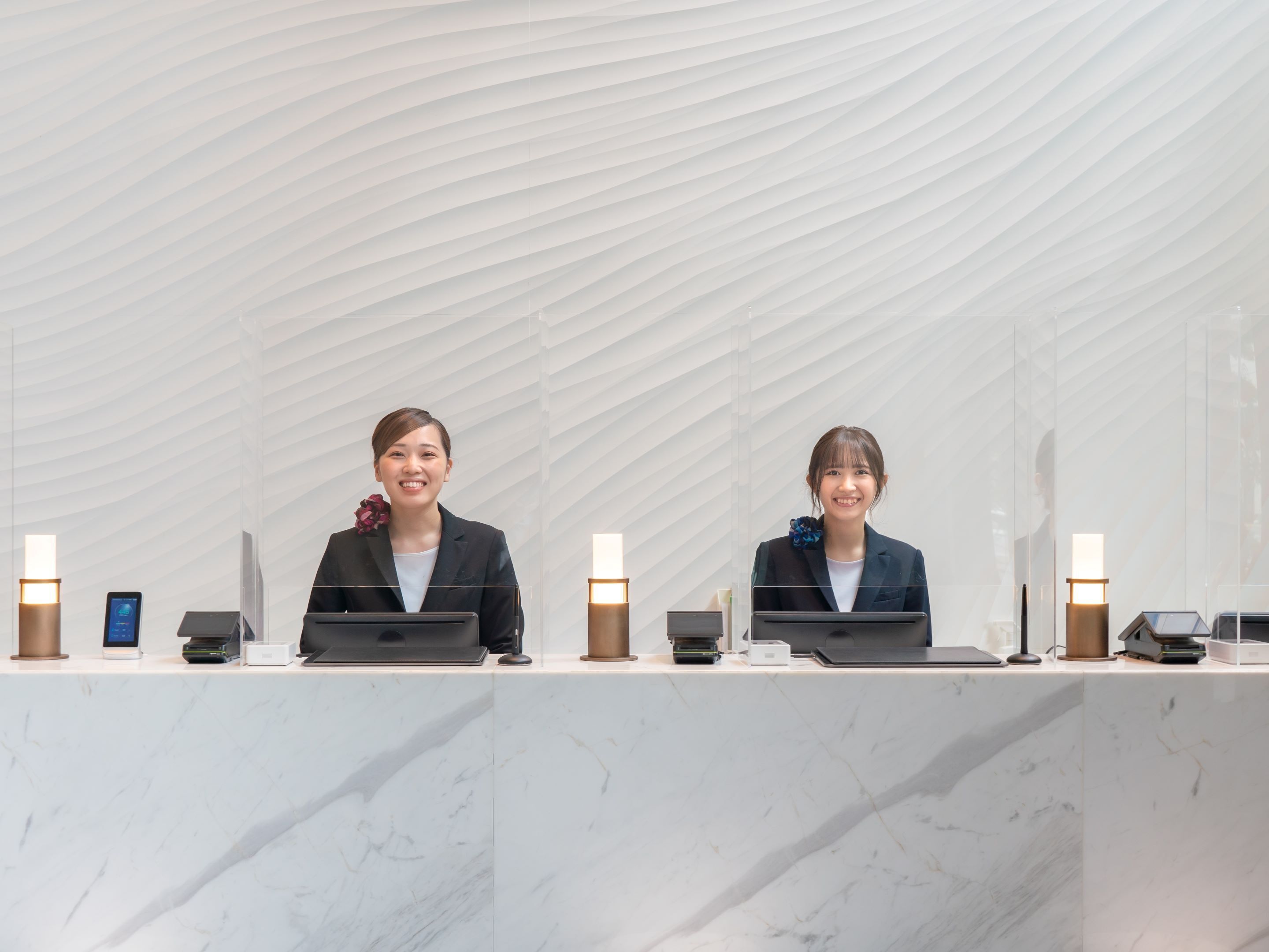 Two receptionists at a reception desk with laptops, a phone, and a tablet.