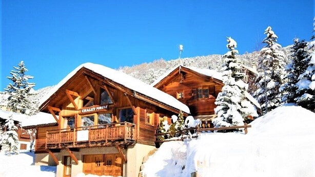 Eine Holzchalet mit Balkon, umgeben von schneebedeckten Bäumen und blauem Himmel.