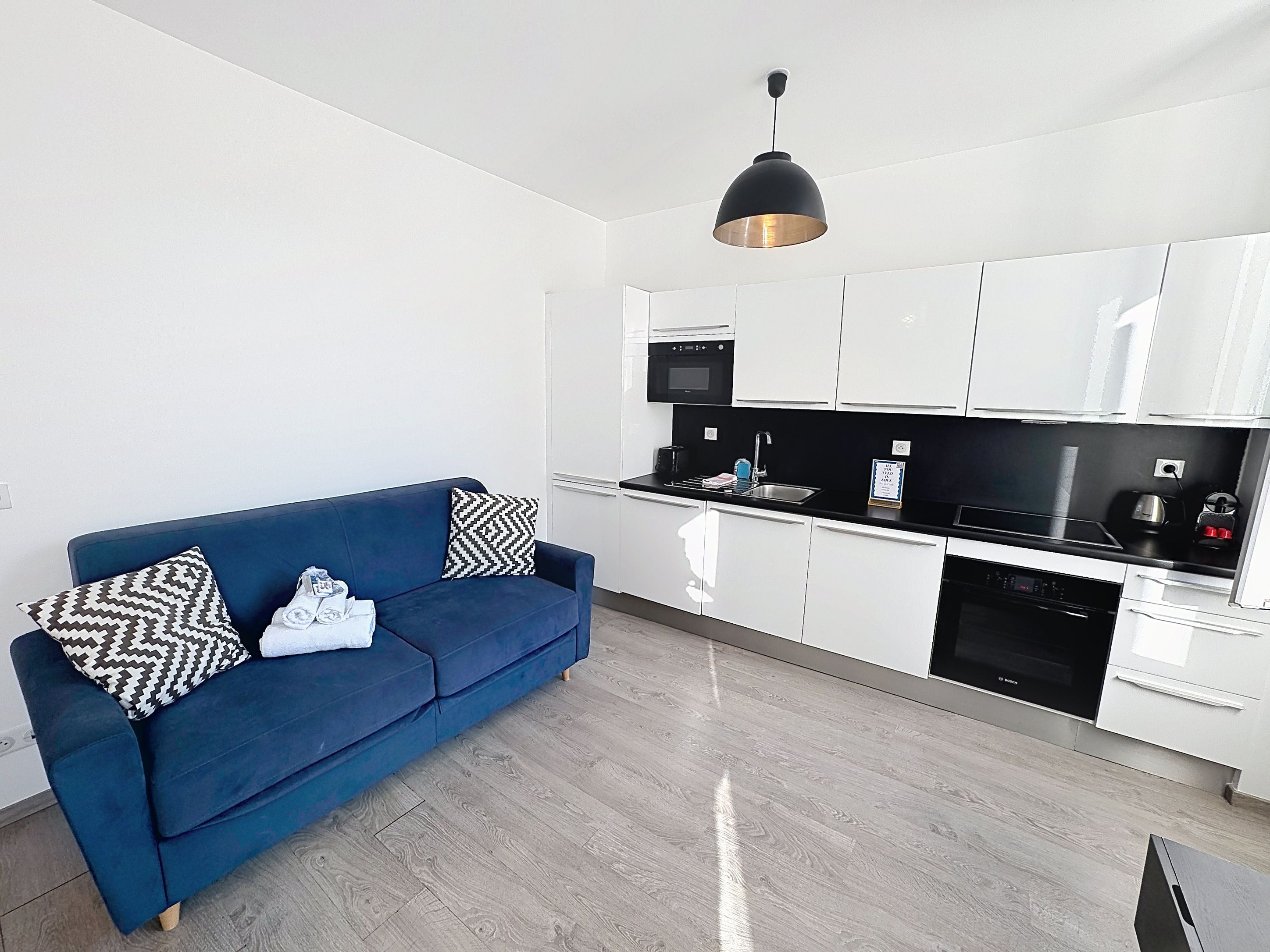 A modern kitchen with white cabinets, a blue sofa with patterned cushions, and a black pendant light.