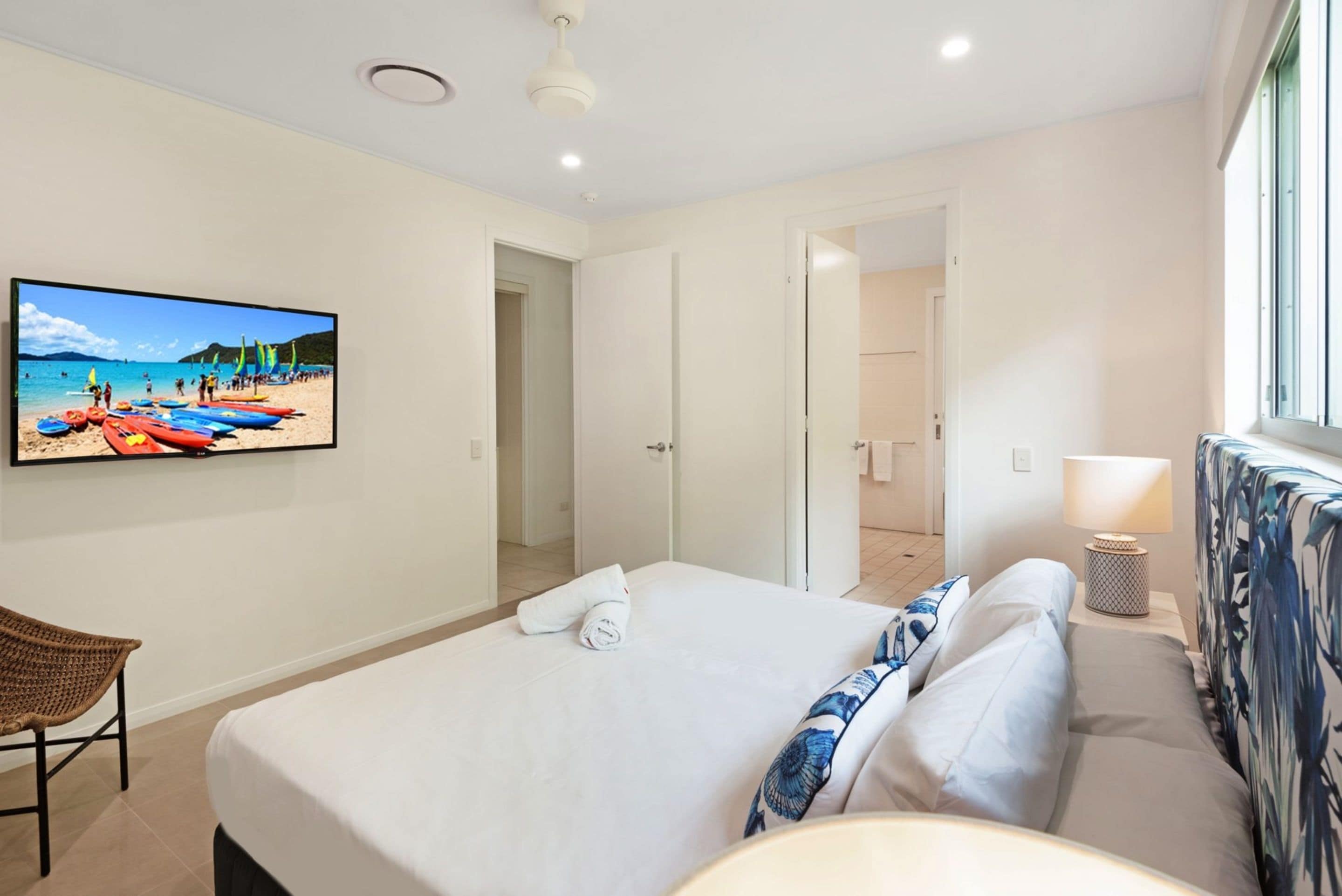 Una habitación de hotel con cama, una televisión mostrando una escena de playa, una ventana con cortinas y una lámpara de noche.